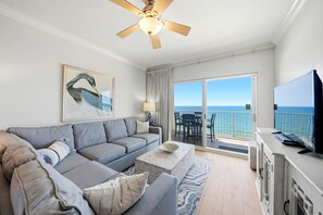 Un soggiorno con divano, tavolino, ventilatore a soffitto, televisore e un balcone con vista sull'oceano.