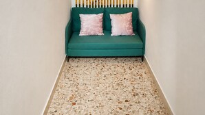 Ein smaragdgrünes Sofa mit rosa Kissen, ein Terrazzo-Boden, eine Wand mit vertikalen Jalousien und Wandleuchten.