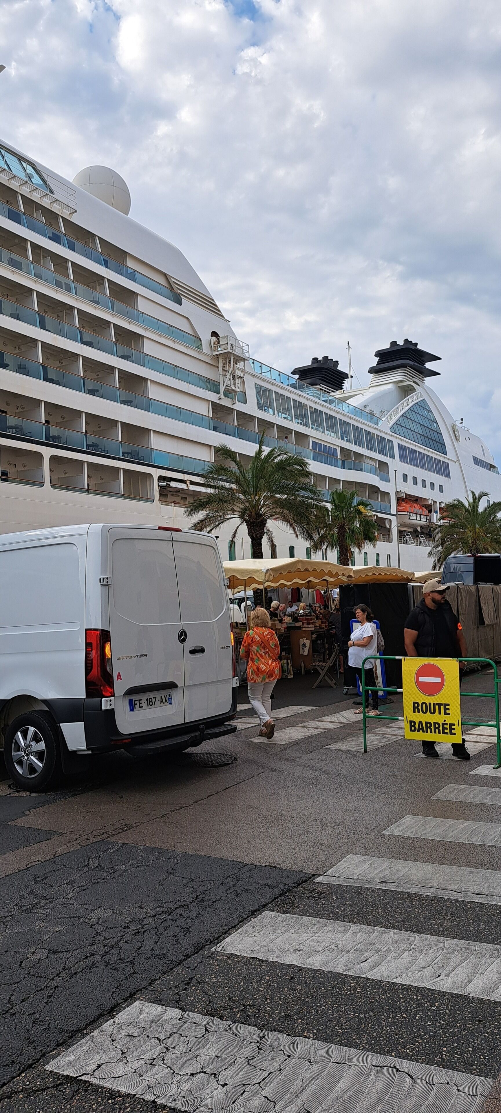 Een cruiseschip met meerdere dekken en balkons, een witte bestelwagen geparkeerd ervoor en een voetgangersoversteek met een 'verboden toegang'-bord.