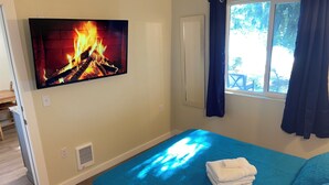 A bedroom with a TV displaying a fireplace, a bed with a blue bedspread, a window with curtains, and a view of the outdoors. - Vacation Home at Lake Serene (Lynnwood)