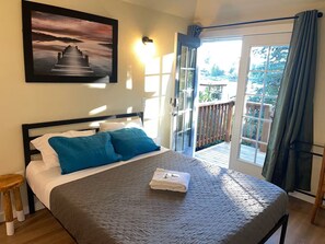 A bedroom with a bed, pillows, a folded towel, a nightstand, a wooden stool, a painting, and a balcony with a view. - Vacation Home at Lake Serene (Lynnwood)