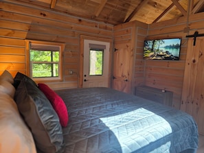 A wooden cabin room with a bed, a door, a window, and a television on the wall.