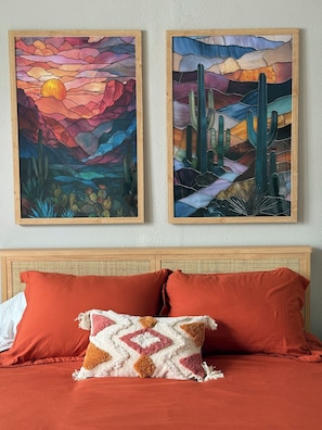 Ein Bett mit roter Bettwäsche und einem gemusterten Zierkissen, ein Holz-Kopfbrett und zwei gerahmte Gemälde, die Wüstenlandschaften zeigen.