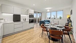 Uma cozinha moderna com armários brancos, sala de jantar com mesa redonda e cadeiras, e um sofá azul ao fundo.