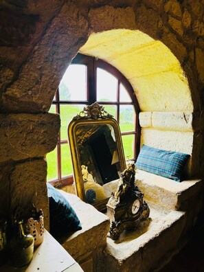 Ein Zimmer mit Steinwänden, einem großen Bogenfenster, einem Spiegel, einer Uhr und einem blauen Kissen auf einer Ablagefläche.