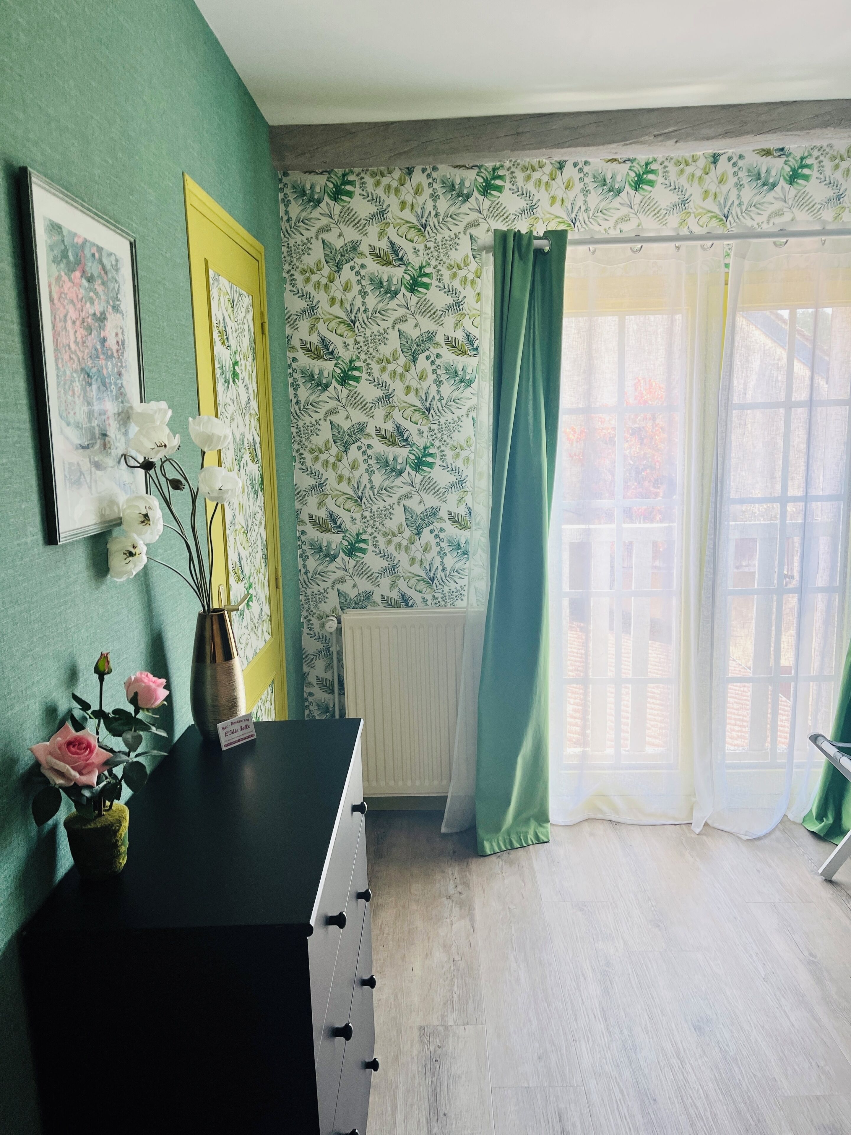 A room with a radiator, a black dresser, a vase with flowers, and a window with curtains.