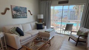 Sala de estar com sofá, poltronas, mesa de centro de vidro, pintura, luminária e vista para uma piscina e um edifício.