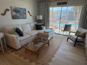 Ein Wohnzimmer mit einer Couch, Sesseln, einem Glastisch, einem Gemälde, einer Lampe sowie Blick auf einen Pool und ein Gebäude.