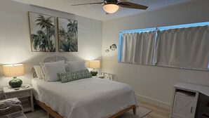 Quarto com cama, duas mesas de cabeceira, escrivaninha, ventoinha no teto e janela com cortinas.