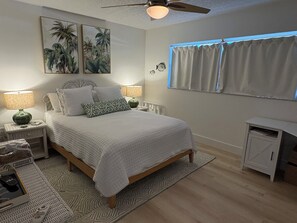 Ein Schlafzimmer mit einem Bett, zwei Nachttischen, einem Schreibtisch, einem Deckenventilator und einem Fenster mit Vorhängen.