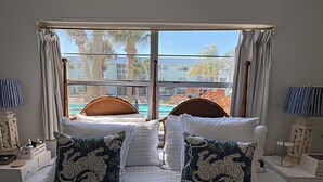 Uma cama de casal com vista para a piscina e palmeiras, duas almofadas decorativas, uma mesa de cabeceira com abajur e uma janela com cortinas.