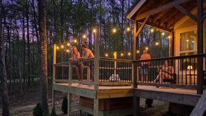 A wooden deck with string lights, a covered seating area, and a small table, and a view of the surrounding forest.
