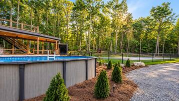 A modern outdoor pool area with a wooden deck and a covered seating area, surrounded by a landscaped garden with pine trees and a gravel path.