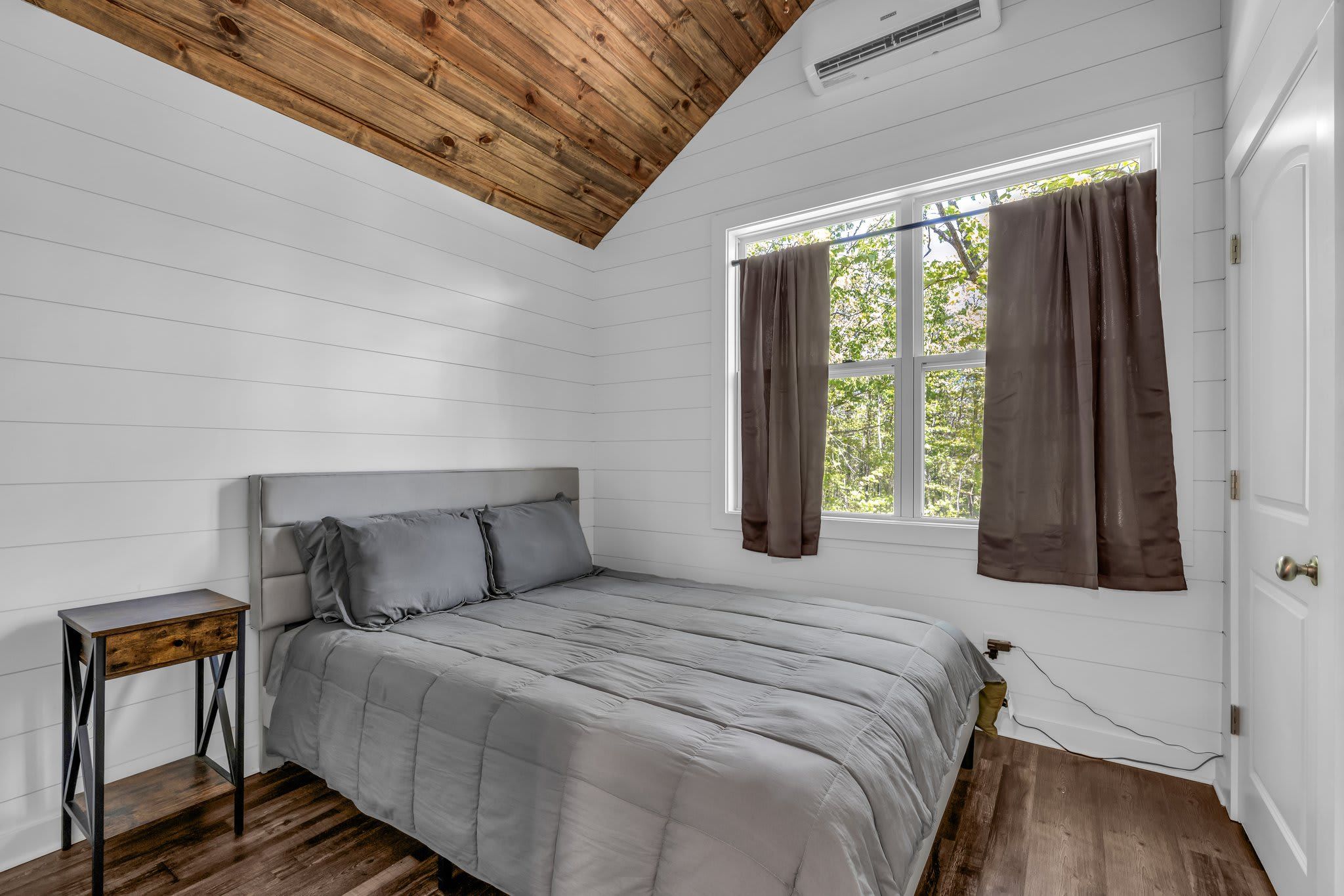 Un dormitorio con una cama, una mesita de noche, un ventanal con cortinas y un techo de madera.