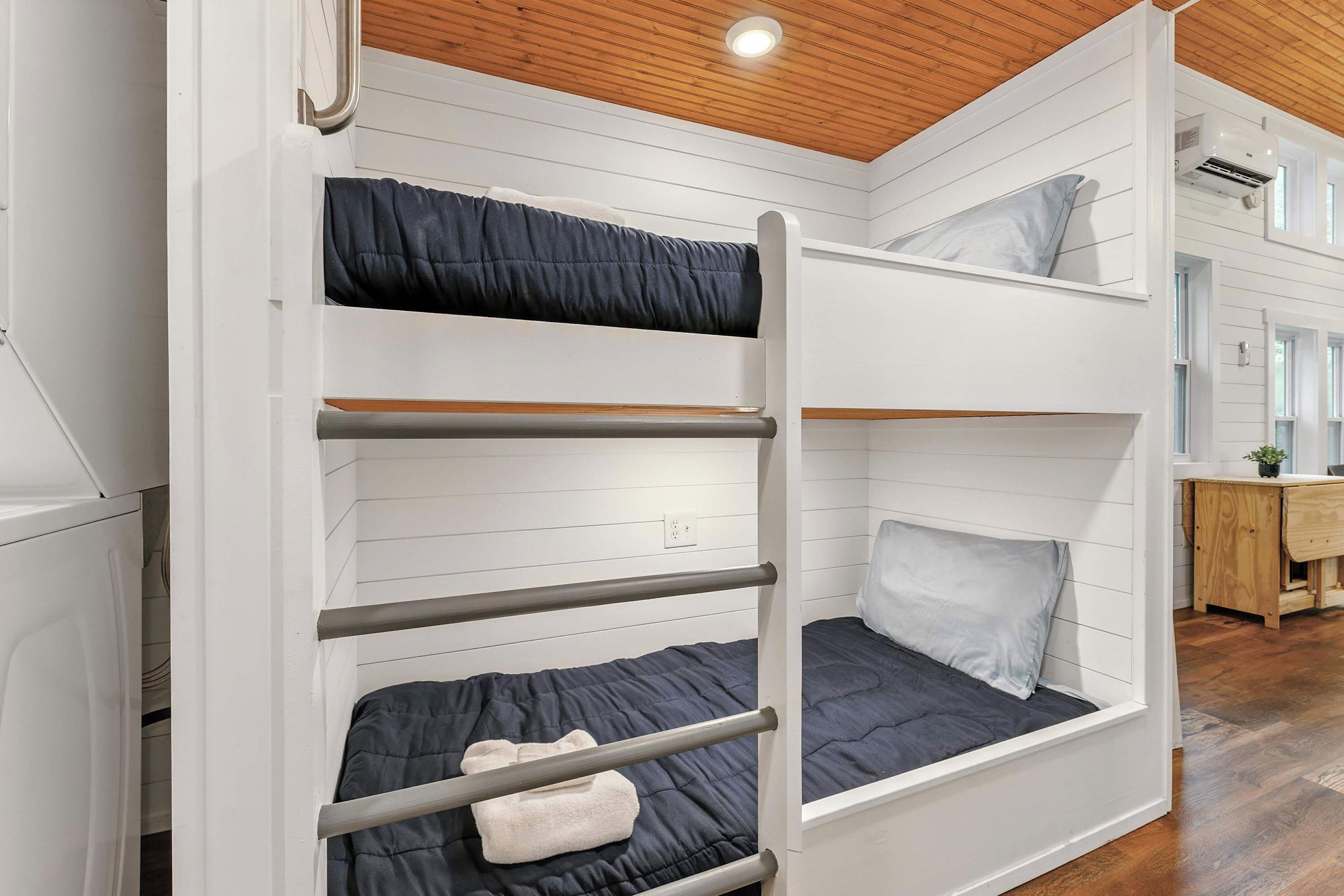 A bunk bed with a wooden ladder, a desk with a chair, and a window in a room with white walls and wooden flooring.