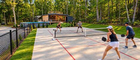 An outdoor tennis court with a wooden fence, a covered seating area, and trees in the background.