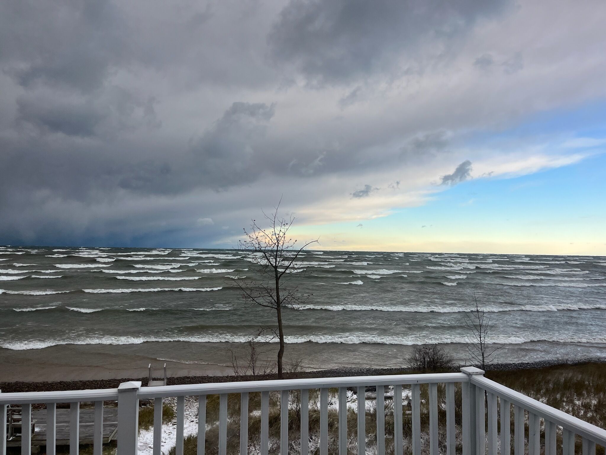 A view of the ocean with waves crashing onto the shore, a cloudy sky, and a wooden deck in the foreground.