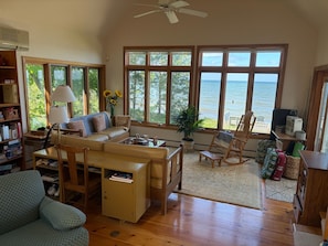 A spacious living room with a large window overlooking the sea, a cozy seating area, and a bookshelf filled with books. - Stunning 4BR Ontario Lake House (Mannsville)