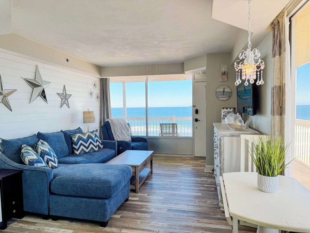 Una sala de estar con un sofá azul, una mesita y vista al mar a través de amplios ventanales.