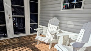 A covered porch with wooden flooring, white Adirondack chairs, and a window with a decorative item. - Turtleheart-Cozy 3BR 2BA in the heart of Oak Island, relax and enjoy island vibe (Oak Island)