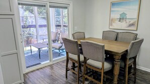 A dining room with a wooden table and six upholstered chairs, a sliding glass door leading to an outdoor patio, and a framed picture on the wall. - Turtleheart-Cozy 3BR 2BA in the heart of Oak Island, relax and enjoy island vibe (Oak Island)