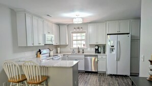 A kitchen with white cabinets, a stainless steel refrigerator, a dishwasher, and a window with a view of greenery. - Turtleheart-Cozy 3BR 2BA in the heart of Oak Island, relax and enjoy island vibe (Oak Island)