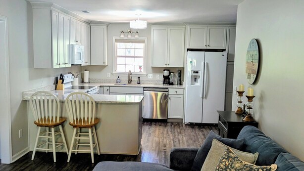 A modern kitchen with white cabinets, a central island, and a dining area with two chairs. - Turtleheart-Cozy 3BR 2BA in the heart of Oak Island, relax and enjoy island vibe (Oak Island)
