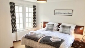 Un dormitorio ordenado con cama, mesitas de noche, lámpara y ventana con cortinas.
