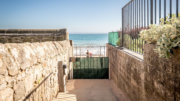 Un muro de piedra que conduce a una puerta con valla metálica, con vista a una playa.