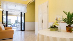 A hotel lobby with a yellow wall, a white door, a round table with a white tablecloth, a yellow armchair, and a potted plant. - Hotel Maria Luisa (Rimini)