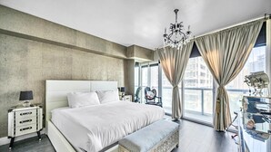 Moderni hotellihuone, jossa on suuri sänky, penkki, yöpöytä, kattokruunu ja suuret ikkunat, joista avautuu näkymä kaupunkiin.