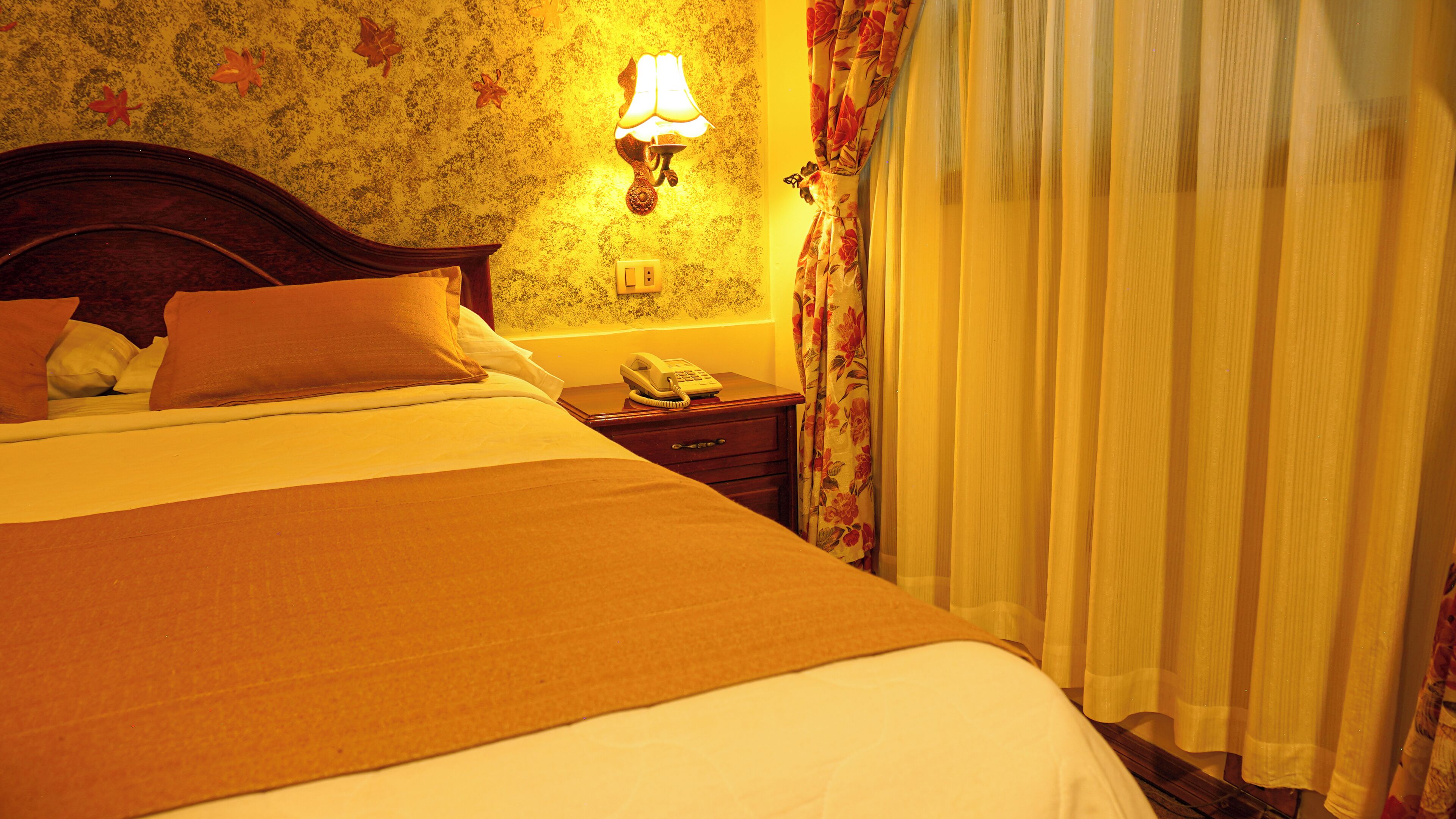 A hotel room with a bed, a bedside table with a lamp, a telephone, and floral curtains.