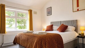 Een hotelkamer met een bed, een raam met gordijnen, een radiator, een aan de muur hangend schilderij en een nachtkastje met een lamp.