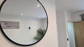 Een kamer met een grote ronde spiegel, een aan de muur hangend kunstwerk, een potplant en een houten deur.