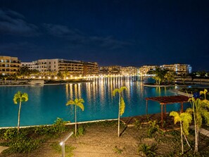 A nighttime view of a resort with a large swimming pool, palm trees, and illuminated buildings in the background.