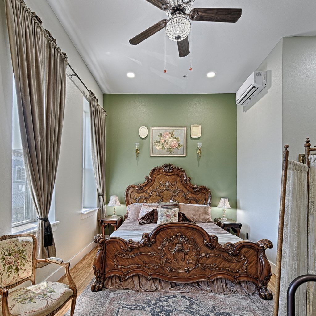 A bedroom with a large bed, a ceiling fan, a window with curtains, a wall-mounted air conditioner, and a floral painting.