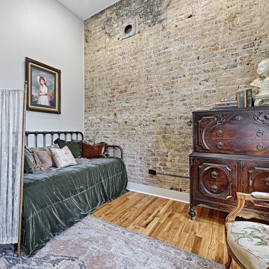 A bedroom with a brick wall, a bed, a dresser, and a chair.