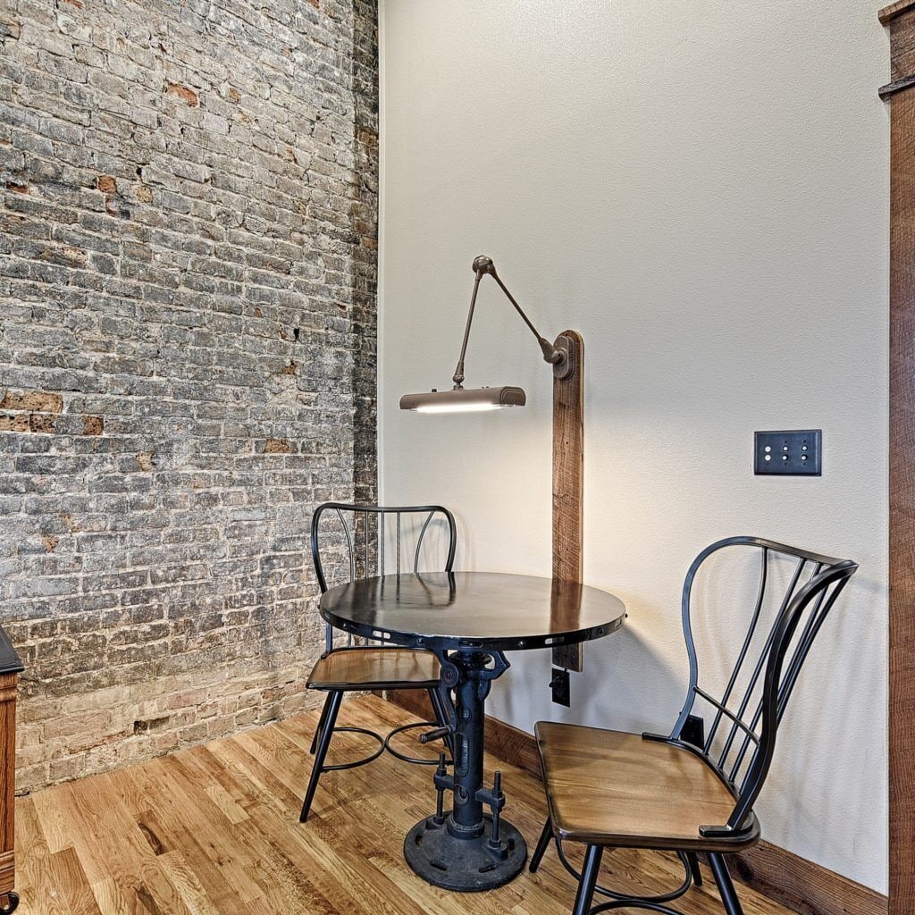 A room with a brick wall, a round table with chairs, a floor lamp, and a power outlet on the wall.