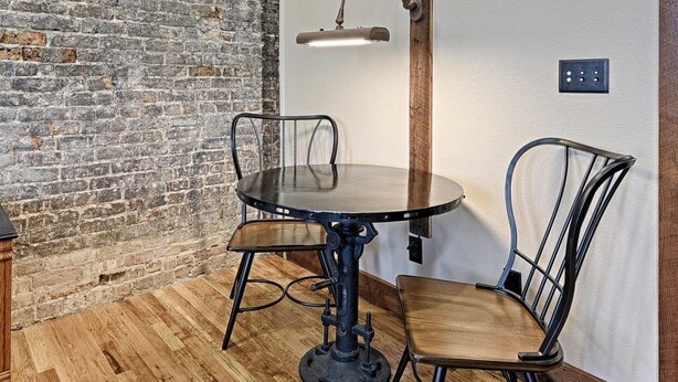 A room with a brick wall, a round table with chairs, a floor lamp, and a power outlet on the wall. - Lofts on Lee (Greenville)