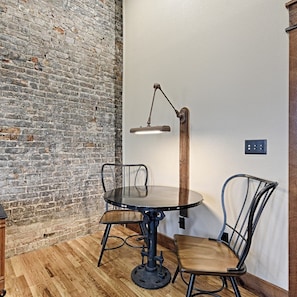 A room with a brick wall, a round table with chairs, a floor lamp, and a power outlet on the wall.