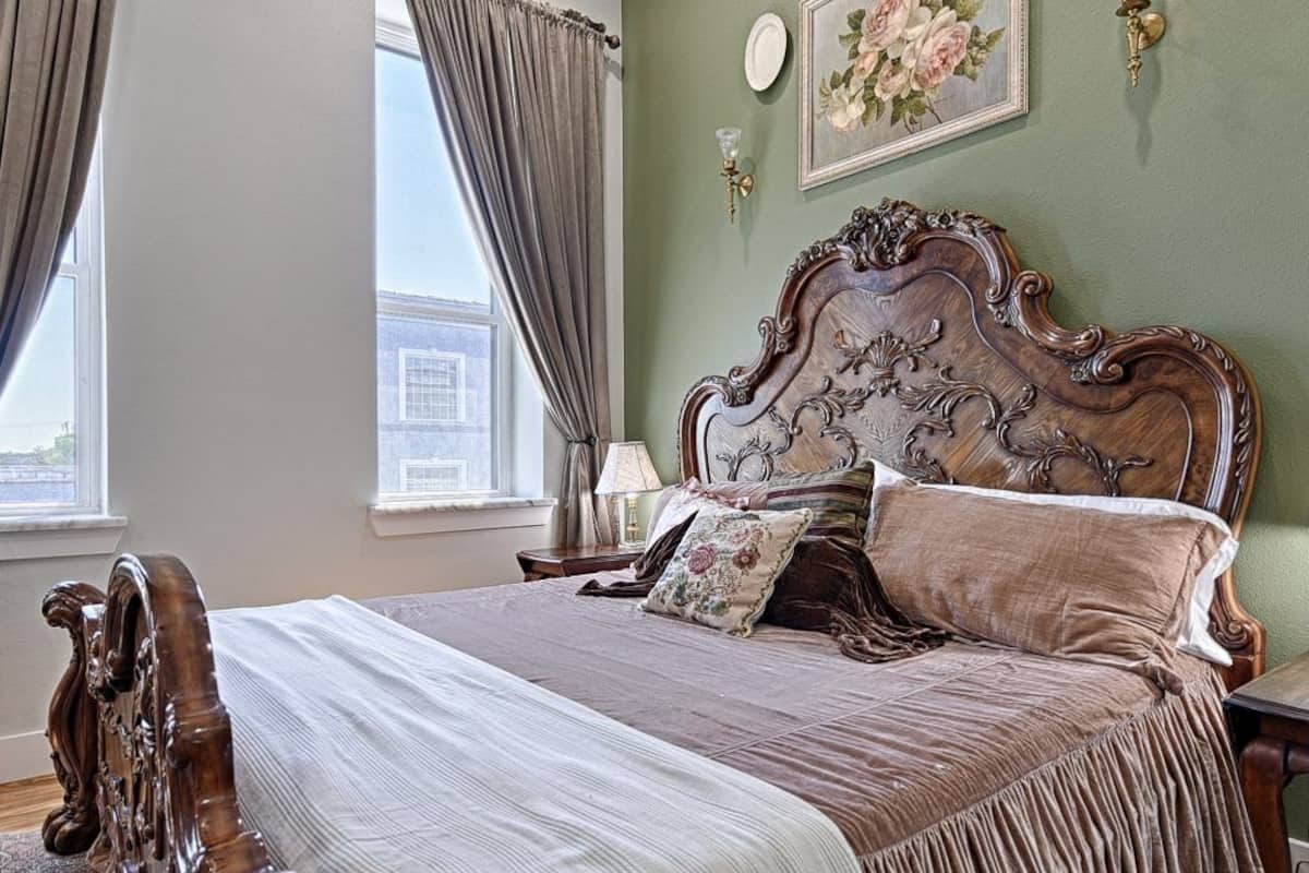 Een slaapkamer met een groot bed, een houten hoofdeinde, een nachtkastje, een lamp, een raam met gordijnen en een bloemendecoratie aan de muur.