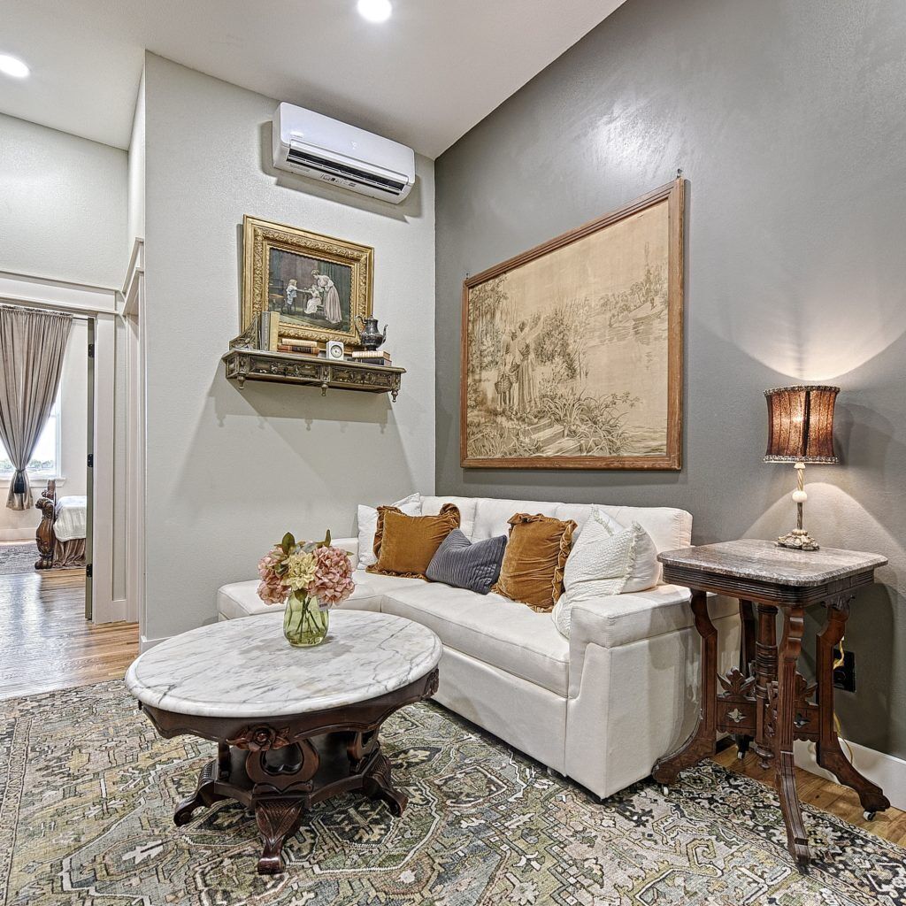 Uma sala de estar com um sofá branco, uma mesa de centro redonda, um tapete estampado, um quadro na parede e um abajur sobre uma mesinha lateral.