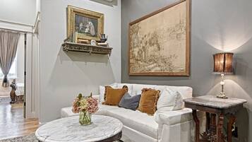 Uma sala de estar com um sofá branco, uma mesa de centro redonda, um tapete estampado, um quadro na parede e um abajur sobre uma mesinha lateral.
