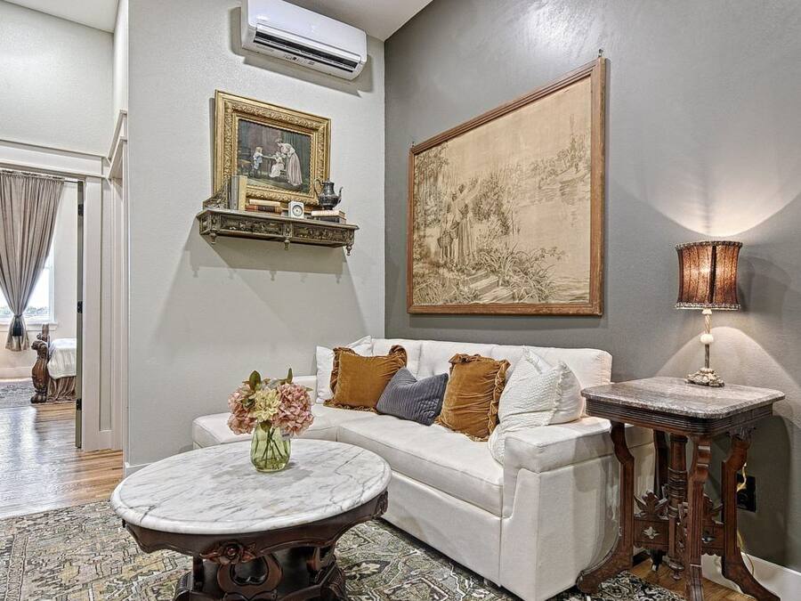 Uma sala de estar com um sofá branco, uma mesa de centro redonda, um tapete estampado, um quadro na parede e um abajur sobre uma mesinha lateral.