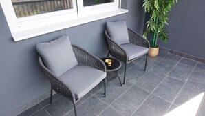Two wicker chairs with grey cushions, a small round table with a gold bowl, and a potted plant on a tiled patio. - Cozy 1-bedroom building apartment in welcoming Kostajnica with AC (Kostajnica)