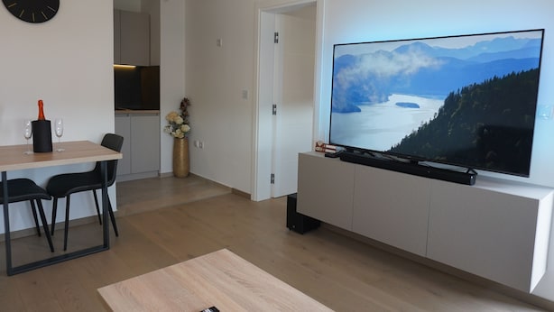 A modern living room with a flat-screen TV, a dining area with a table and chairs, a kitchen in the background, and a door leading to another room. - Cozy 1-bedroom building apartment in welcoming Kostajnica with AC (Kostajnica)