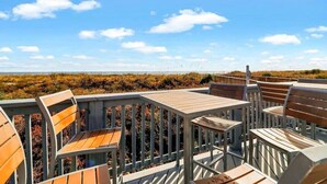A deck with wooden chairs and tables, overlooking a beach with dunes and a clear sky. - Modern Ocean Oasis - Private Beach • Pool • OceanViews (Hilton Head Island)