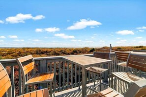 Eine Terrasse mit Holzkubeln und Tischen, mit Blick auf einen Strand mit Dünen und blauem Himmel.