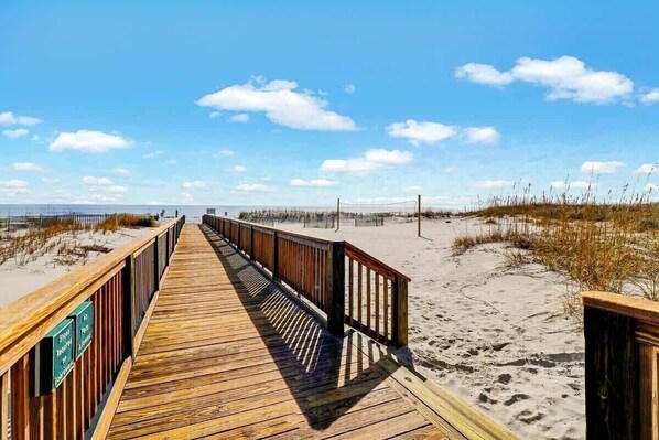 Ein hölzerner Steg führt zu einem Strand mit Dünen und blauem Himmel.