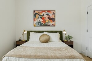 A neatly made bed with a beige blanket, a green upholstered headboard, and a colorful abstract painting above it. - Cozy and Modern 1-bedroom house in Edmond (Edmond)